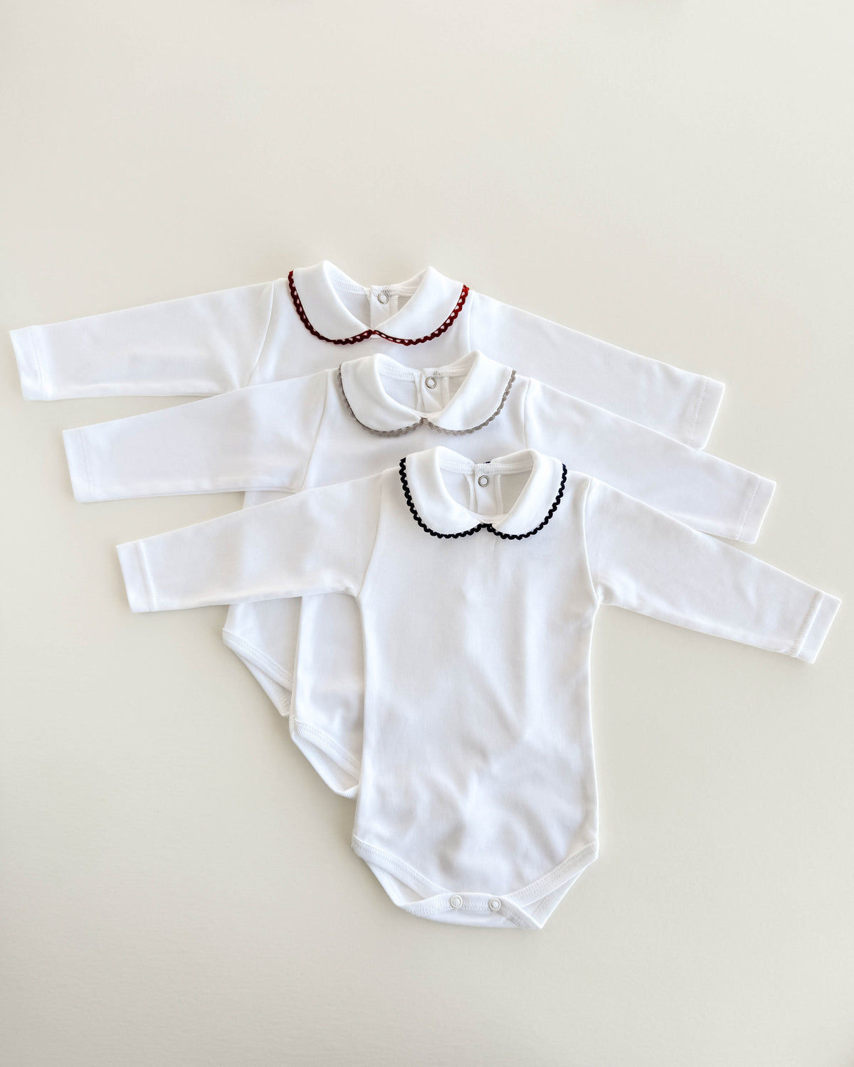 Three white long-sleeve baby bodysuits with Peter Pan collars are arranged in a fan shape on a light beige background. The top bodysuit has a red beaded collar, the middle has a silver beaded collar, and the bottom has a black beaded collar.