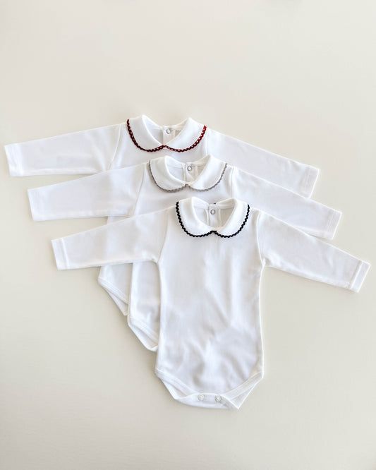 Three white long-sleeve baby bodysuits with Peter Pan collars are arranged in a fan shape on a light beige background. The top bodysuit has a red beaded collar, the middle has a silver beaded collar, and the bottom has a black beaded collar.